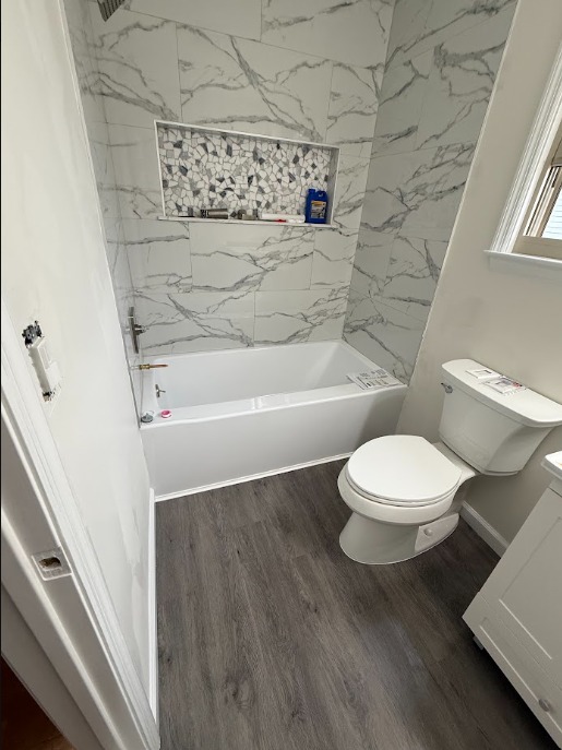 Full bathroom view with a gray marble-look tub surround, mosaic recessed niche, wood-look floor, and bright natural light